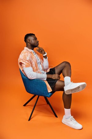 Fashionable African American man sitting in a stylish chair against a bright orange background.
