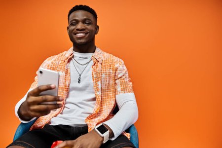Photo for Handsome African American man in stylish attire sitting in a chair while using a cell phone. - Royalty Free Image