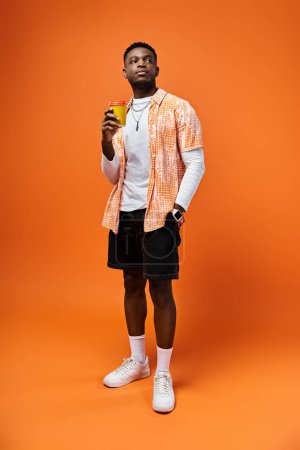 Foto de Hombre afroamericano guapo con atuendo de moda sosteniendo una bebida frente a un fondo naranja brillante. - Imagen libre de derechos