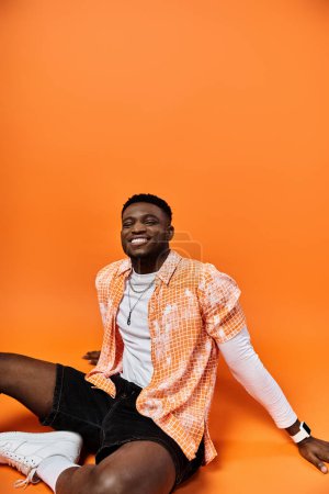 Fashionable African American man in orange shirt relaxing on bright orange background.