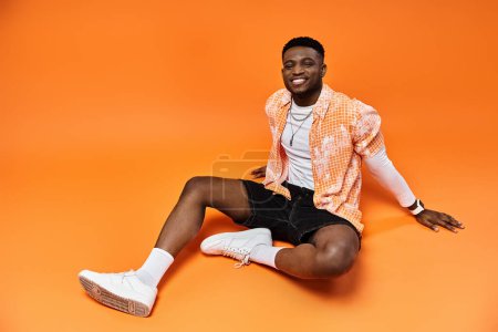 Foto de Fashionable young African American man in stylish attire sitting on bold orange backdrop. - Imagen libre de derechos