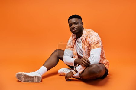 Schöner junger afroamerikanischer Mann in stylischem orangefarbenem Hemd sitzt anmutig auf dem Boden.