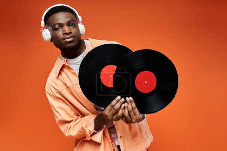 Handsome African American man in stylish attire holding vinyl record on orange background.