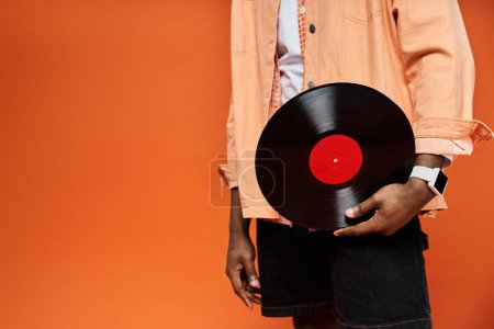 Fashionable young African American man in stylish attire holding a vinyl record against an orange backdrop.