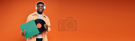 Photo for Young African American man in fashionable attire holding a vinyl record in orange backdrop. - Royalty Free Image