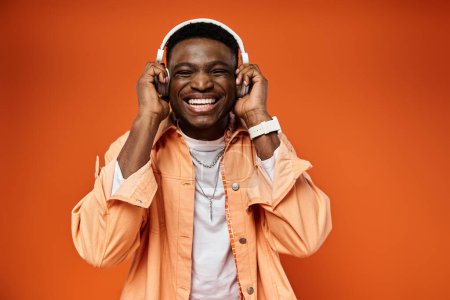 Foto de Elegante hombre negro sonríe alegremente mientras usa auriculares contra el vibrante telón de fondo naranja. - Imagen libre de derechos