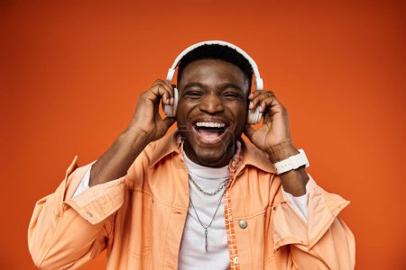 Stylish black man listening to music with headphones on bright orange background.