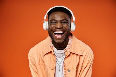 Stylish black man smiles wearing headphones on orange background.