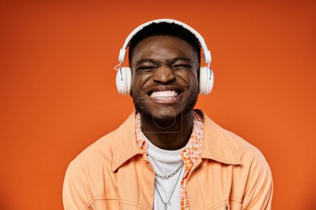 Foto de Elegante hombre afroamericano con auriculares sonríe contra fondo naranja. - Imagen libre de derechos