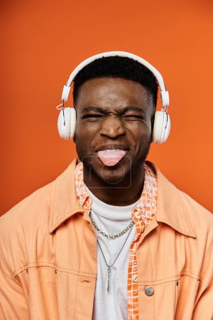 Téléchargez les photos : Homme afro-américain élégant portant des écouteurs avec la langue qui sort dans un geste amusant et ludique. - en image libre de droit
