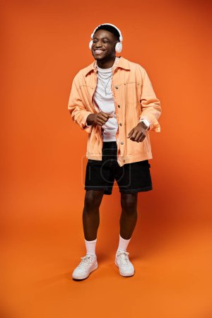 Ein modischer junger Afroamerikaner in orangefarbener Jacke und Shorts posiert selbstbewusst vor einem dazu passenden orangefarbenen Hintergrund.
