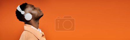Foto de Young African American man in white attire standing in front of vivid orange background with white headphones. - Imagen libre de derechos