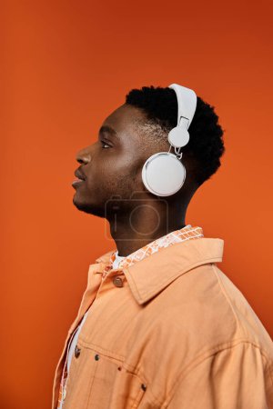 Jeune homme afro-américain à la mode appréciant la musique avec des écouteurs devant un fond orange vif.