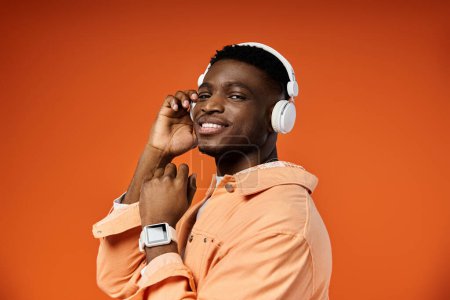Foto de Cool, hombre negro joven con estilo posando con auriculares contra el fondo naranja vibrante. - Imagen libre de derechos
