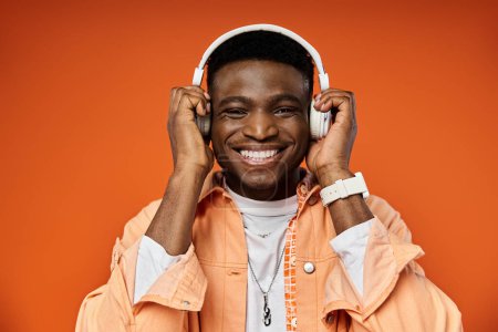Téléchargez les photos : Beau Afro-Américain en tenue élégante, souriant tout en portant des écouteurs sur fond orange. - en image libre de droit