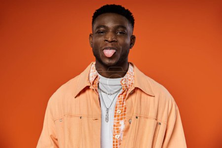 A fashionable, young African American man sticks out his tongue while wearing a stylish orange jacket.