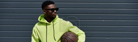 Schöner afroamerikanischer Mann in grünem Kapuzenpullover hält Basketball.
