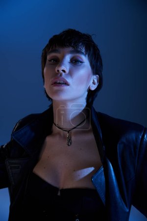 Foto de Stylish woman in black leather jacket striking a pose under a blue light. - Imagen libre de derechos