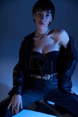 Stylish young woman poses confidently in leather jacket on blue background.