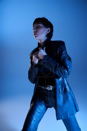 Foto de Young woman strikes a pose in leather jacket against blue backdrop. - Imagen libre de derechos
