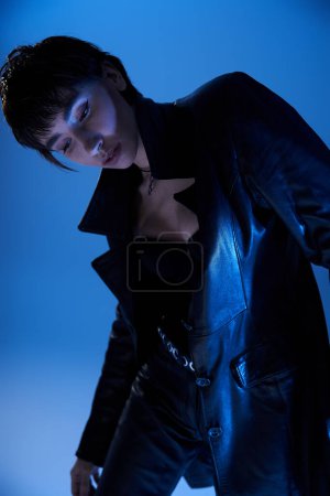 Foto de A young woman in a black leather jacket striking a pose against a blue background. - Imagen libre de derechos