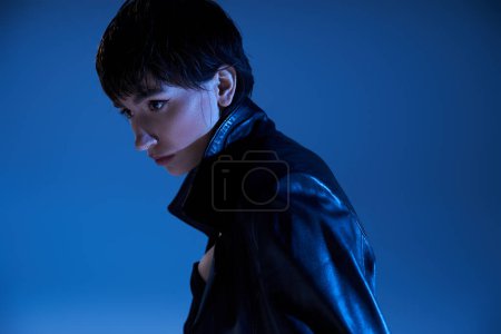Foto de Trendy young woman in leather jacket poses confidently in front of a vivid blue background. - Imagen libre de derechos
