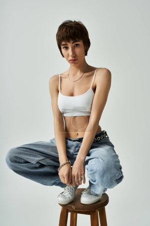 Téléchargez les photos : Young woman in white top, jeans, posing on stool. - en image libre de droit