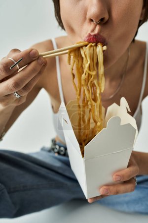 A beautiful young woman in fashionable attire eating noodles directly from a box.