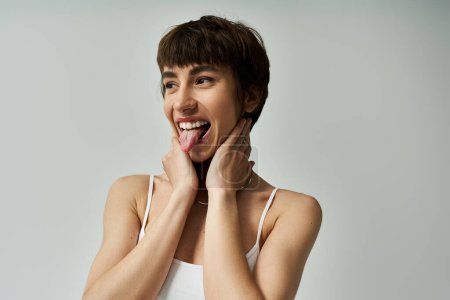 Young woman in stylish attire playfully sticking out tongue against white background.