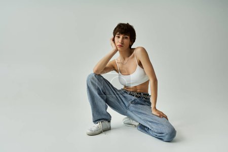 A stylish woman in a white tank top and jeans sitting gracefully on a white background.