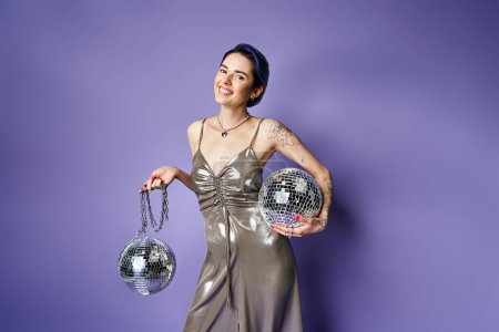 A stylish woman in a silver dress holding two disco balls, radiating glamour and fun in a studio setting.