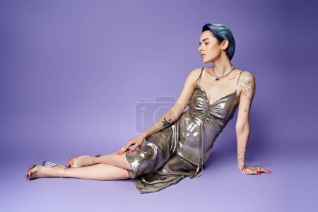 Young woman with short blue hair, in silver party dress, sitting gracefully on the floor in a studio setting.