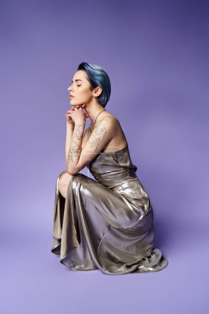 A young woman with blue hair is seated on a purple background, striking a pose in a silver party dress.