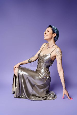 Young woman with short blue hair striking a pose while seated on the ground in a stunning silver dress.