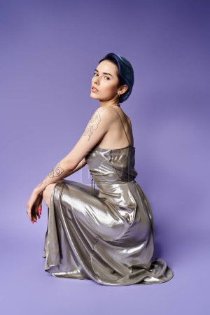 A stunning young woman with short blue hair sits gracefully in a shimmering silver dress against a vibrant purple backdrop.