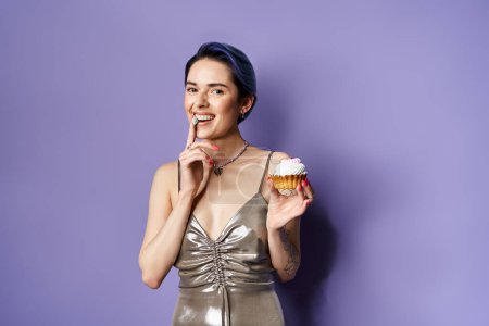 Téléchargez les photos : A stylish young woman with short blue hair elegantly holds a cupcake in a silver party dress in a studio setting. - en image libre de droit