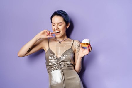 A young woman with short blue hair elegantly holds a cupcake, showcasing a silver party dress in a studio setting.