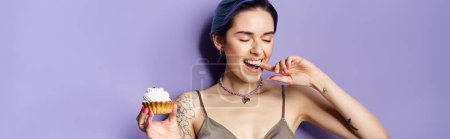 A pretty young woman with short blue hair in a silver party dress joyfully holds and savors a delicious cupcake.