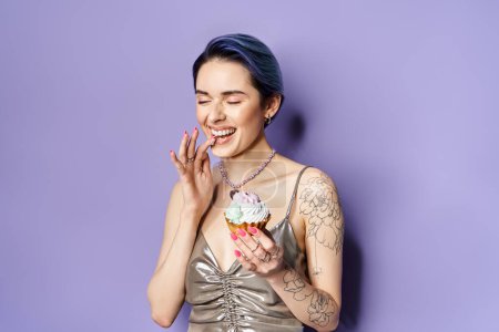 A stylish young woman with blue hair holds a beautifully decorated cupcake in a dazzling silver dress.