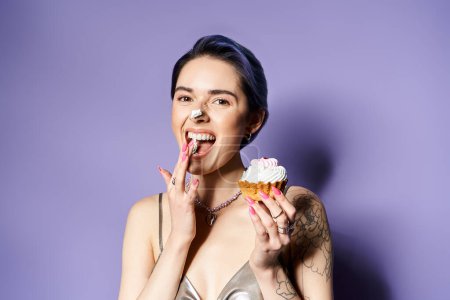 Téléchargez les photos : Young woman with short blue hair holding cupcake in front of mouth, dressed in silver party attire. - en image libre de droit