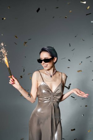 Young woman in a silver dress with blue hair holding a sparkler in a stylish and festive pose.