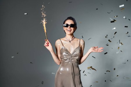 Young woman with blue hair poses in silver dress, holding sparkler.