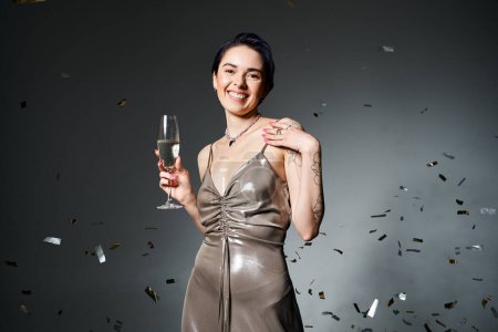 A young woman with short blue hair in a silver party dress holds a glass of champagne in a studio setting.