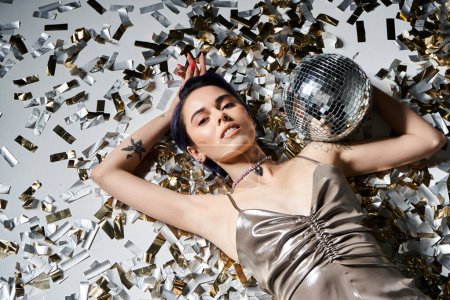 A stylish young woman with blue hair dazzles in a silver dress while holding a reflective disco ball.