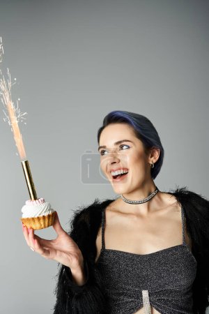 Young woman in stylish attire holding cupcake with lit sparkler in hand, celebrating a special occasion.