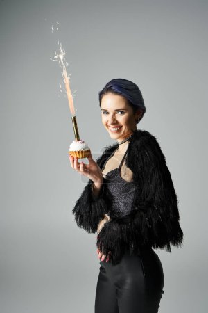 A young woman with short, dyed hair posing in stylish attire, holding a cupcake with a sparkler.
