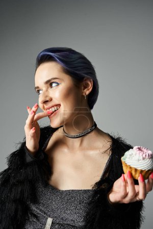 A stylish young woman with blue hair joyfully holds a cupcake.