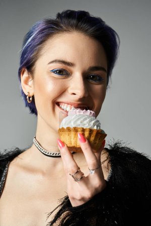A pretty young woman with blue hair joyfully eating a cupcake in a studio setting, celebrating like a birthday girl.