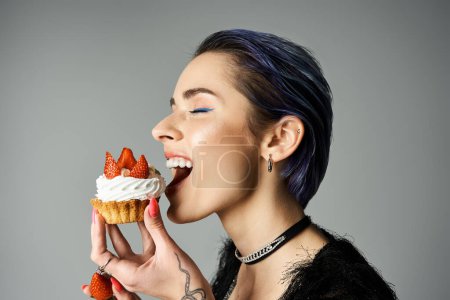 A young woman with short dyed hair smiling while eating a cupcake topped with strawberries.