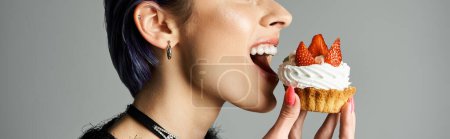 A young woman joyfully eats a cupcake topped with icing and strawberries in a stylish studio setting.